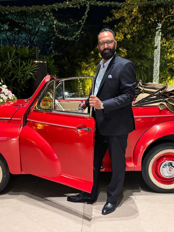 Govind Raj in a suit beside a red classic convertible